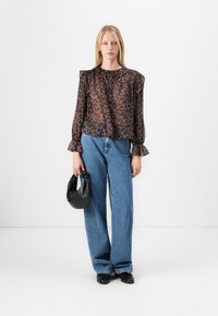 Blusa nera trasparente con motivo floreale, spalle arricciate e scollo con bottoni, abbinata a jeans a gamba larga blu chiaro e una borsa nera.
