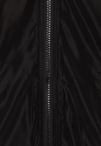Veste noire zippée en nylon, d'une texture lisse, avec un accent en métal argenté au centre. Design minimaliste sans impressions supplémentaires.