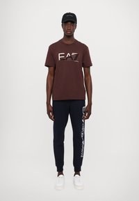 Burgundi pamut póló "EA7" logóval, sötétkék melegítőnadrággal, amelyen fehér felirat található az oldalon. Fehér sportcipőkkel egészíti ki az öltözéket.