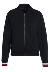 Námořnicky modrá bunda na zip vyrobená z jemné látky, s vysokým límcem, dvěma bočními kapsami a žebrovanými manžetami s červenými a bílými pruhy.