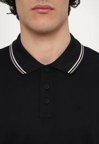 Tricou polo negru cu guler clasic, decorat cu dungi albe și bej, cinci nasturi negri și un logo subtil pe partea stângă.