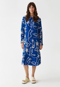 Vestito blu con bottoni e motivi astratti bianchi, maniche lunghe e colletto. Abbinato a sandali con plateau di colore chiaro.