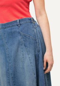 Mujer vistiendo una falda de mezclilla azul de tiro alto con cremallera lateral y una blusa sin mangas de color rojo brillante, con la mano descansando a su lado.