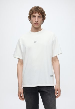 Junger Mann mit lockigem Haar, der ein schlichtes weißes T-Shirt mit kurzen Ärmeln und kleinem Logo auf der Brust sowie dunkle Jeans trägt, steht vor einem weißen Hintergrund.