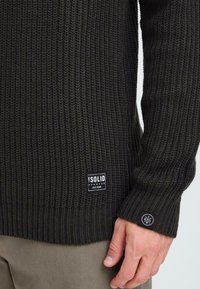 Dunkelgrüner Strickpullover mit gerippter Textur, einem Logo-Patch am Saum und einem runden Logo-Akzent am Bündchen.