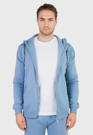 Man met een lichte huid en bruin haar, gekleed in een lichtblauwe hoodie met ritssluiting over een wit T-shirt en bijpassende lichtblauwe broek.