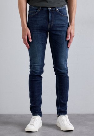 Jean en denim bleu foncé, skinny-fit, avec une texture lisse, comportant cinq poches, des coutures visibles et une fermeture à bouton en laiton. Porté avec des baskets blanches.