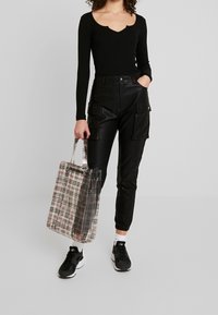 Černý dlouhý rukáv top s výstřihem do vodorovného tvaru, v kombinaci s černými kalhotami z umělé kůže s kapsami po stranách a průhlednou taškou s kostkovaným vzorem.