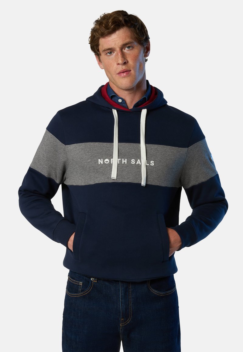 Hoodie color-block navy et gris avec cordons blancs et logo. Comprend une poche kangourou et des poignets côtelés. Texture de tissu douce.