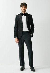 Czarna aksamitna marynarka smokingowa z satynowymi klapami, zestawiona z czarnymi spodniami i białą koszulą. Akcesoria obejmują czarny muszkieter oraz buty.