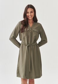 Olive green dress with long sleeves, button-down front, two chest pockets, and a tied waist belt. Smooth fabric with a classic silhouette.