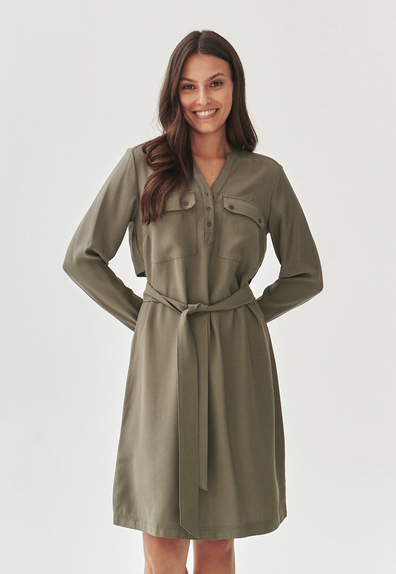 Olive green dress with long sleeves, button-down front, two chest pockets, and a tied waist belt. Smooth fabric with a classic silhouette.