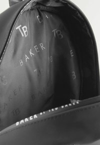 Unutrašnjost crnog ruksaka s ponavljajućim tekstom "BAKER" i "TB" u suptilnom, sjajnom printu na mat crnoj tkanini.