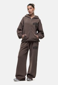 Brauner Zip-Hoodie und passende Sweatpants aus weichem Stoff. Mit Fronttaschen und bedrucktem Text "A GOOD NOTHING" auf beiden Teilen.