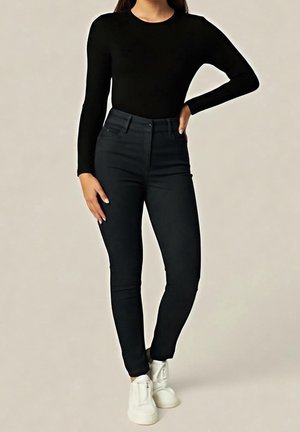 Frau trägt schwarzen Langarmtop und hoch taillierte schwarze Skinny Jeans mit weißen Sneakers, steht vor einem schlichten hellen Hintergrund.