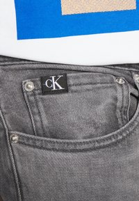 Närbild av en ficka på grå denimjeans med en liten svart "cK"-tyg etikett på övre kanten, under en vit och blå skjortkant.
