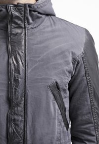 Mörkgrå jacka med en kombination av matt tyg och glänsande läderdetaljer, med hög krage och en sidoficka. Texturerad yta synlig.