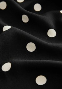 Schwarzer Stoff mit einer glatten Textur, der große, gleichmäßig verteilte weiße Pünktchen aufweist. Die Pünktchen haben einen sanften Glanz und eine leichte Formvariation.