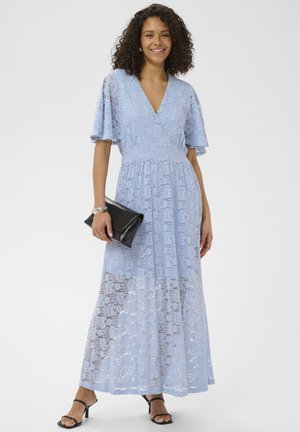 Femme debout et souriante, portant une robe longue en dentelle bleu clair, des sandales noires à talons, tenant une pochette noire.