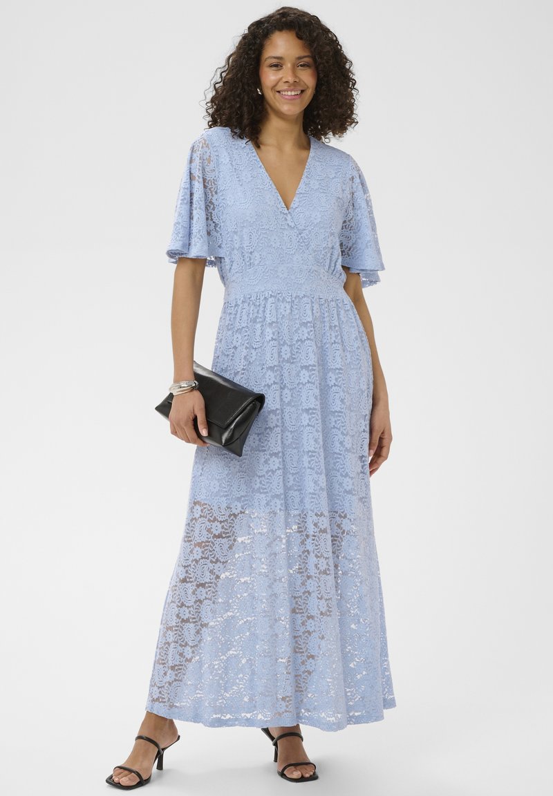 Femme debout et souriante, portant une robe longue en dentelle bleu clair, des sandales noires à talons, tenant une pochette noire.