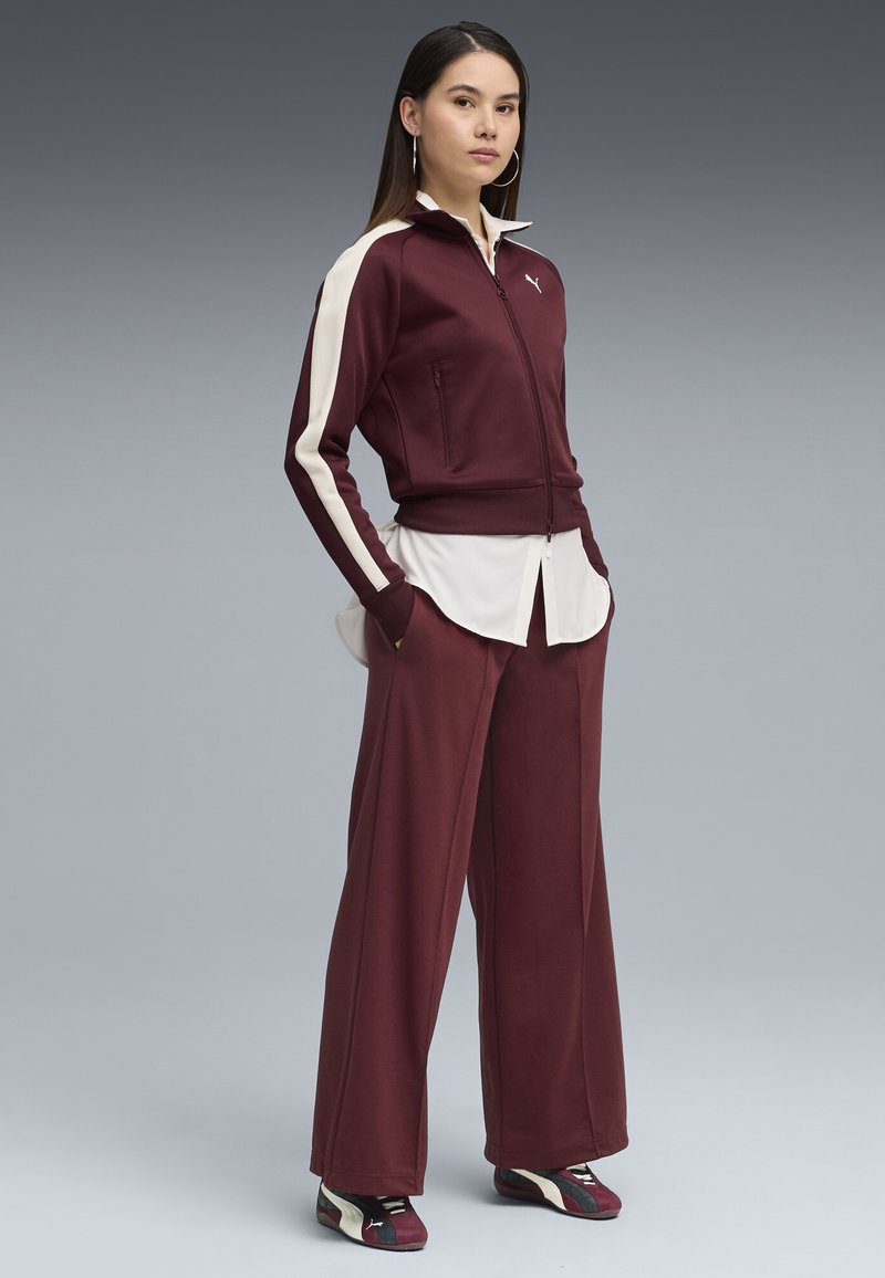 Tuta bordeaux con strisce a contrasto crema, giacca con zip, pantaloni larghi e calzature con accenti neri. Tessuto liscio, vestibilità casual.