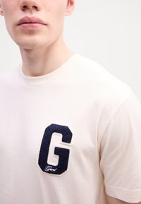 Gros plan sur une personne portant un t-shirt blanc avec un grand patch en lettre « G » texturé bleu marine sur la poitrine.