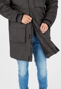 Veste matelassée gris foncé avec une fermeture éclair à l'avant et des accents en Velcro ; dotée de poches latérales et d'une coupe décontractée, associée à un jean bleu clair.