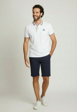 Homme souriant marchant les mains dans les poches, portant un polo blanc, un short bleu marine et des baskets blanches sur un fond uni.