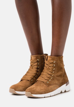 Bottes basses en daim marron avec un design à lacets, œillets dorés et une semelle en caoutchouc blanche présentant un motif texturé. Fermeture éclair sur le côté.