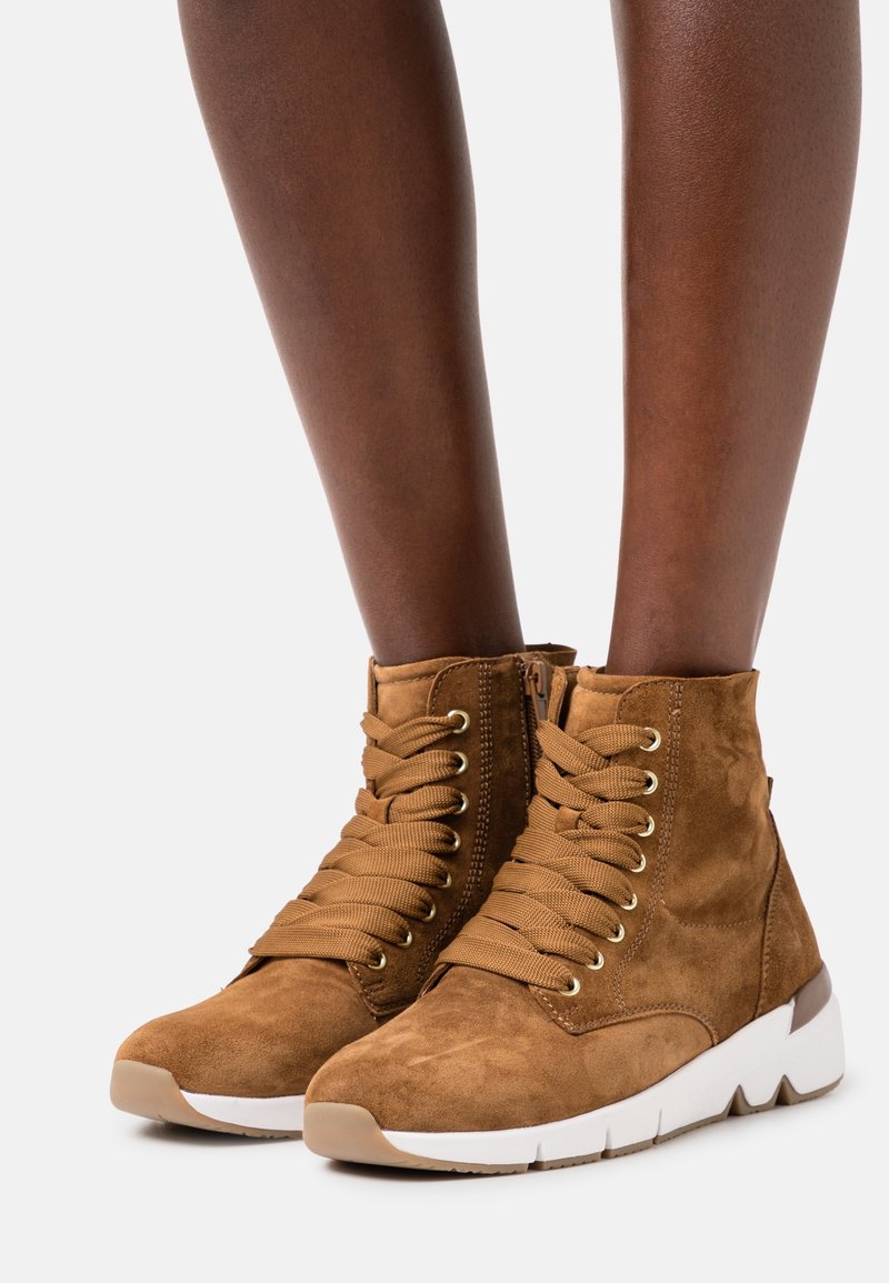 Bottes basses en daim marron avec un design à lacets, œillets dorés et une semelle en caoutchouc blanche présentant un motif texturé. Fermeture éclair sur le côté.