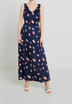 Mouwloze maxi-jurk in marineblauw met rood-witte bloemenprint, V-hals, getailleerde taille, gedragen met witte sandalen met hak.