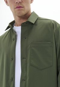 Olivaroheline button-up särk terava kraega, rindadaskuga ja tekstureeritud kangast. Kihtide kaupa valge ümmarguse kaelusega T-särgi peal.