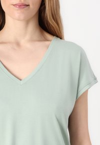 Tricou verde deschis cu guler în V, cu mâneci scurte, realizat dintr-un material fin, având o croială lejeră și detalii de cusătură subtile.