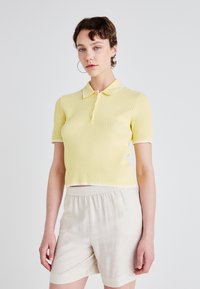 Polo shirt giallo a coste con colletto rigido, maniche corte e cinque bottoni, abbinato a pantaloni corti beige chiaro.
