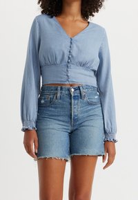 Blusa cropped azzurro chiaro con maniche lunghe a sbuffo e dettagli con bottoni sul davanti, abbinata a pantaloni shorts di denim strappati.