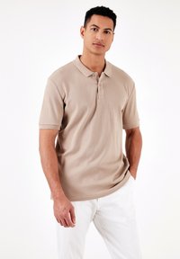 Vaaleanbeige puolihihainen polo, jossa on ribbetty pinta, kolme nappia ja tavallinen kaulus. Yllä vaaleat housut.
