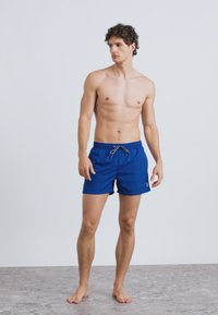 Siniset uimashortsit, jotka on valmistettu kevyestä kankaasta. Niissä on joustava vyötärönauha ja kiristysnauha. Shortsien malli esitellään seisovalla miesmallilla.