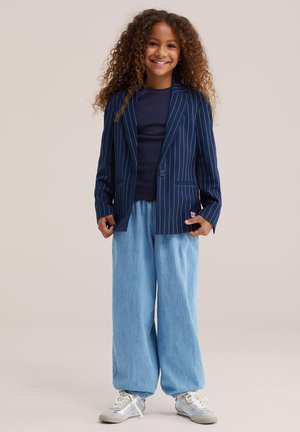 Enfant souriant aux cheveux bouclés portant un blazer bleu marine à fines rayures, une chemise sombre, un pantalon large bleu clair et des baskets blanches, debout sur un fond uni.