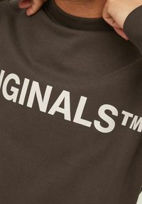 Persona che indossa un sudore marrone con il testo bianco "ORIGINALS™", tenendo il collo vicino al collo con entrambe le mani.