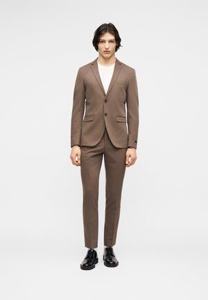 Junger Mann steht frontal in einem maßgeschneiderten braunen Anzug mit weißem Hemd und schwarzen Anzugschuhen vor einem schlichten hellen Hintergrund.