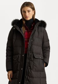 Duga crna puffer jakna s kapuljačom od krzna, koja ima kopče na dugmad, prednje džepove i kviltani dizajn. Nosi se uz crvenu kariranu košulju.