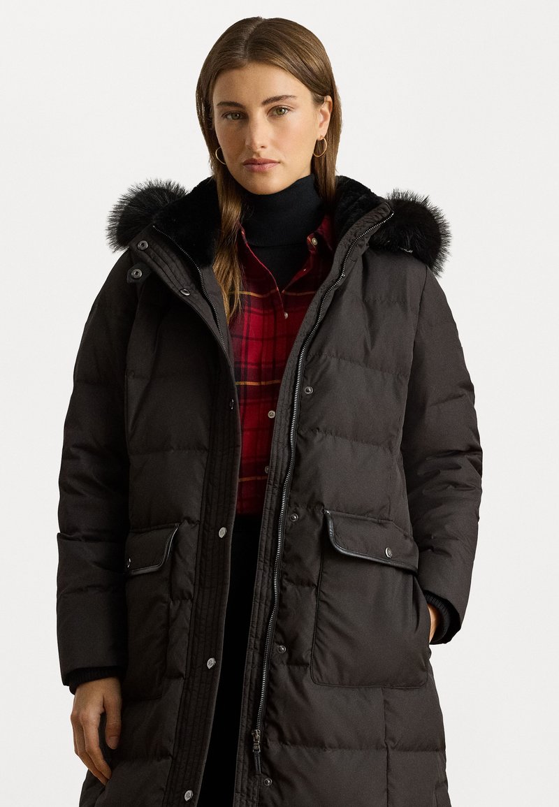 Duga crna puffer jakna s kapuljačom od krzna, koja ima kopče na dugmad, prednje džepove i kviltani dizajn. Nosi se uz crvenu kariranu košulju.