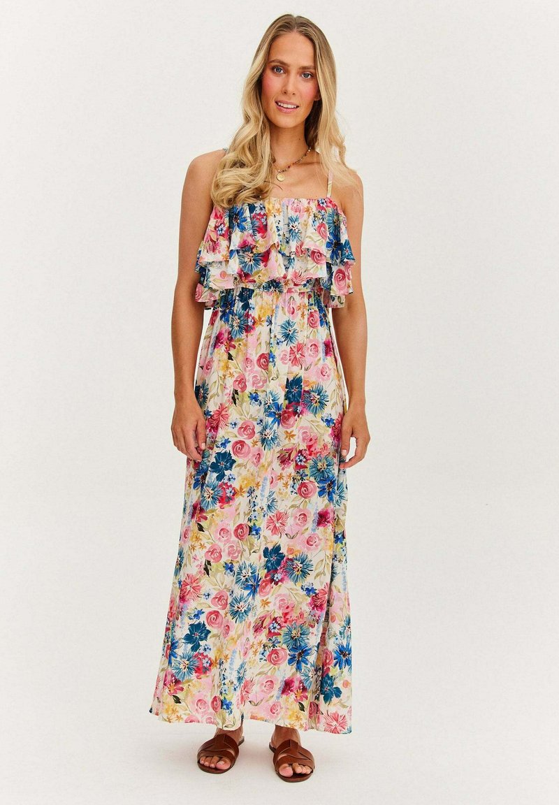 Kwiatowa sukienka maxi z falbaną na górze, z różowymi, niebieskimi i żółtymi kwiatami na jasnym tle. Noszona z brązowymi sandałami.