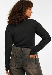 Schwarzes Langarmoberteil aus dehnbarem Stoff, kombiniert mit hochgeschnittenen, ausgewaschenen grauen Jeans mit zwei Rückentaschen.