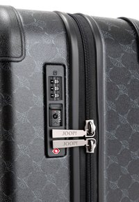 Zwarte koffer met textuur oppervlak, combinatie slot en dubbele ritsen gemarkeerd "JOOP!" met een subtiel bloemenpatroon en een TSA-slot.