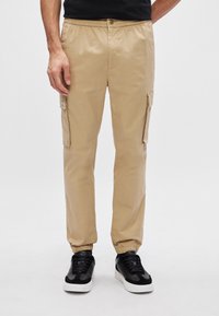 Khaki cargobroek met elastische tailleband en boorden, voorzien van zijzakken en een gestructureerde stof. Gepaard met zwarte sneakers.