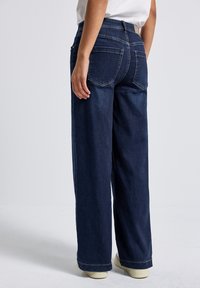 Jeans larges bleu foncé en denim, dotés de poches arrière et d'une taille haute. Le tissu a une texture lisse et des coutures contrastantes.