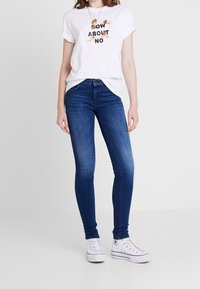 Camiseta blanca de algodón con el texto negro "¿QUÉ TAL NO?", combinada con jeans ajustados azules y zapatillas altas blancas. Tejido suave y ajuste clásico.