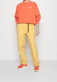 Pomarańczowa bluza z małym logo, w połączeniu z żółtymi spodniami z kordurowej tkaniny ze sznurkiem w pasie oraz czerwonymi trampkami z białymi paskami.