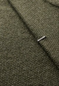 Strukturierter Stoff in dunkelgrün mit einem gewebten Muster. Enthält ein kleines silbernes Hardware-Tag mit der Aufschrift "SUPERDRY".